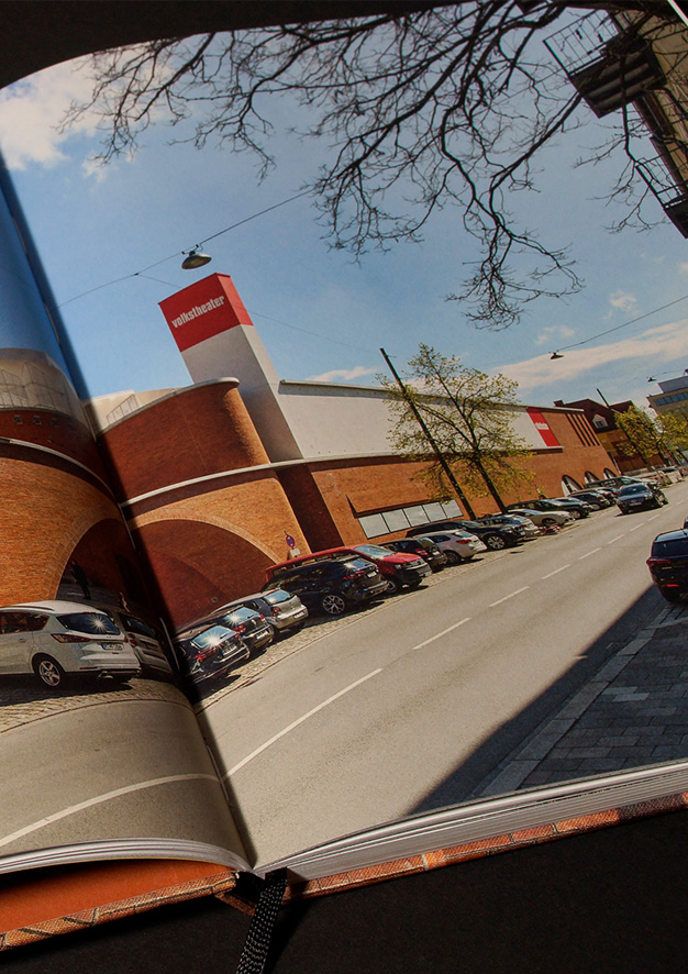 Aufgeschlagene Doppelseite mit schräger Außenansicht entlang der Straße: Vor der Frontfassade eine Reihe parkender Autos.